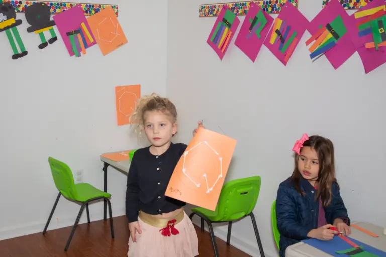 A girl proudly holds up her orange artwork while a friend works at the table behind her