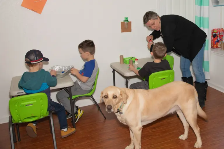 Children work at their tables while a teacher helps and a yellow Labrador wanders by