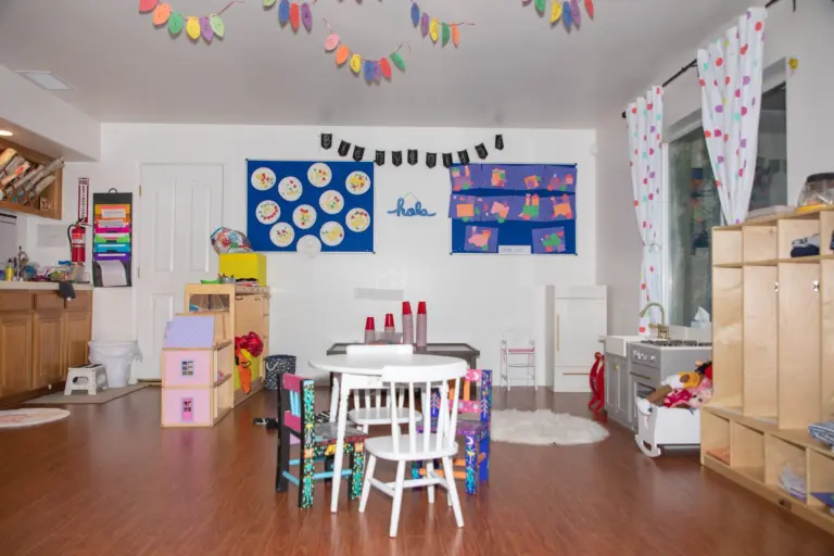 A bright classroom with a white table and chairs, a play kitchen, shelves of toys and a rainbow of bunting