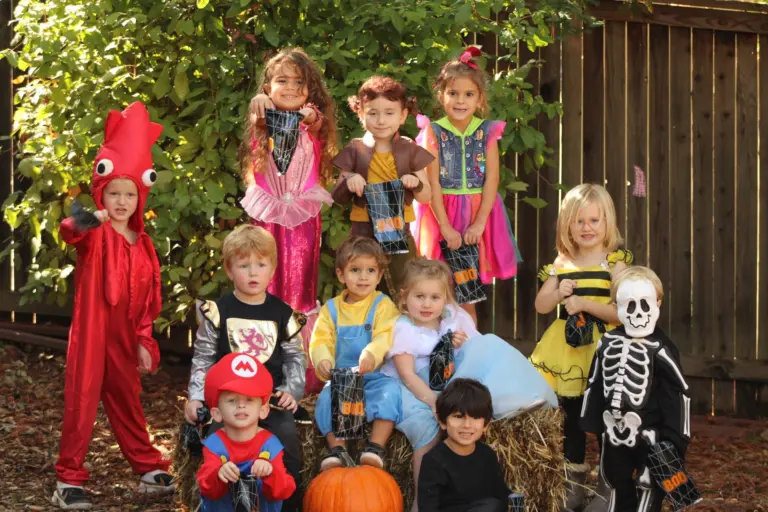 A group of children in Halloween costumes pose together on hay bales with a pumpkin