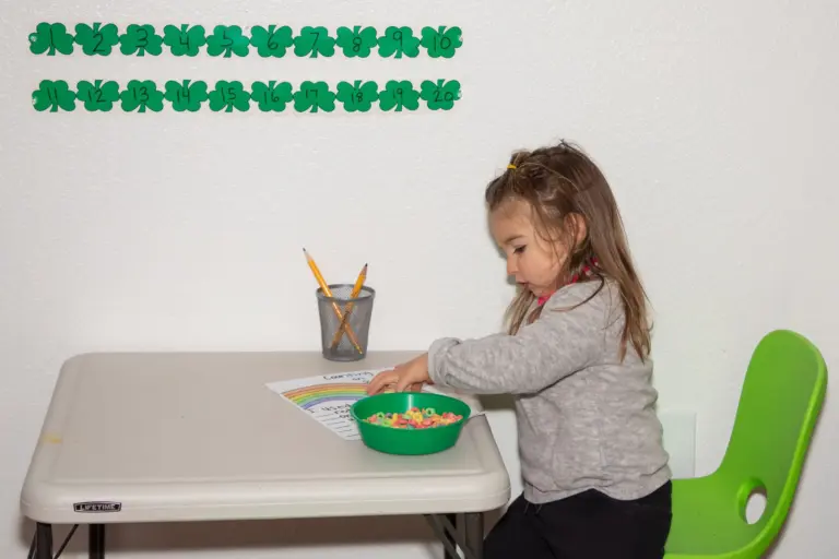 A girl counts small objects from a green bowl at a table under a row of paper shamrocks