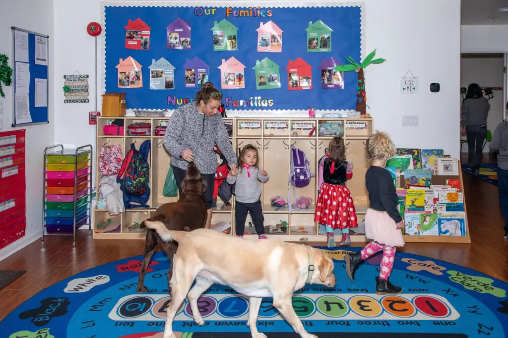 Children hang up their backpacks at the cubbies under the Our Families board while a Labrador trots past