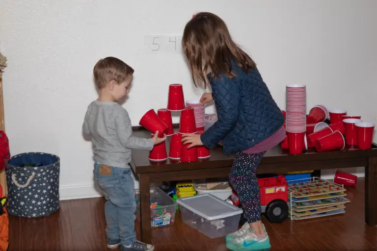 A boy and a girl build towers out of red plastic cups on a low shelf