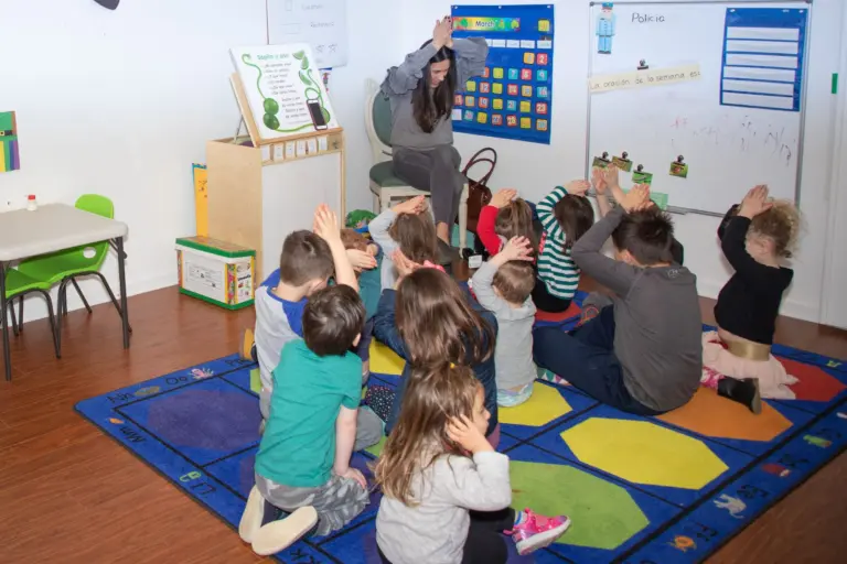 Children on the circle-time rug raise their hands high to answer a question