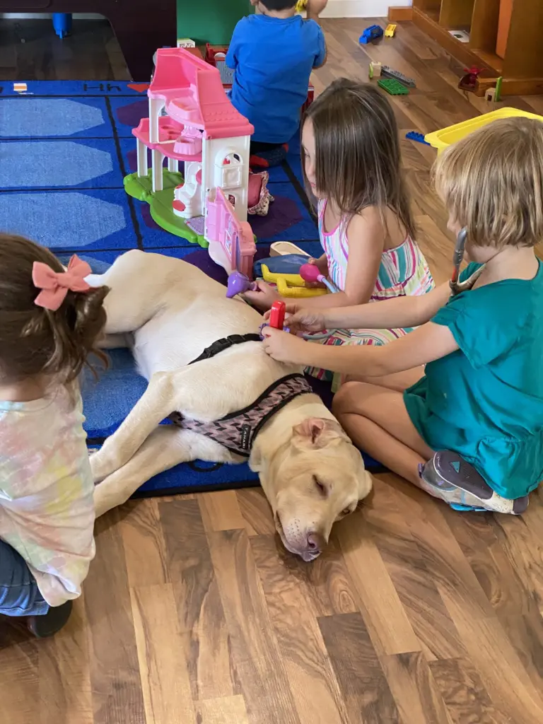 Children sit on the floor petting a calm yellow Labrador beside a pink dollhouse