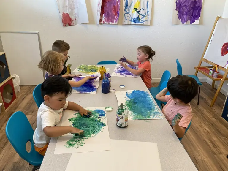 Children paint with bright blues and greens on big sheets of paper around a classroom table