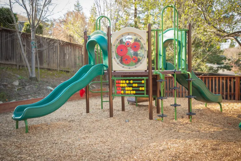 A play structure with a green slide in a wood-chip yard among the trees