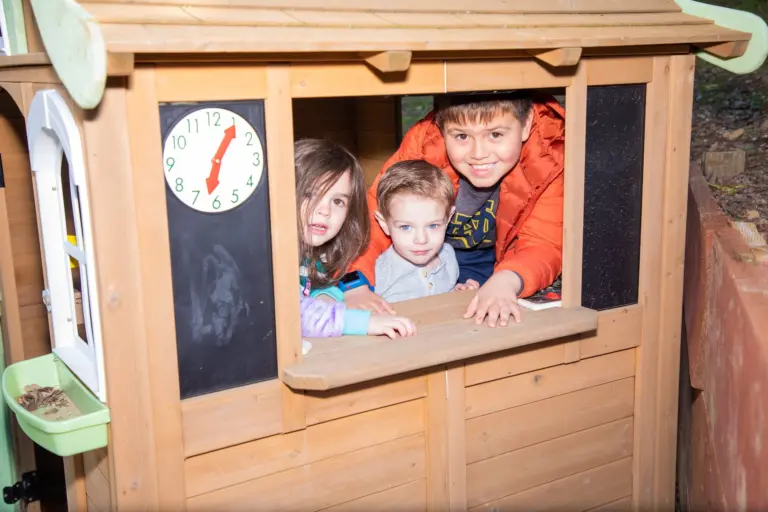Three grinning children peek out of the window of a wooden playhouse
