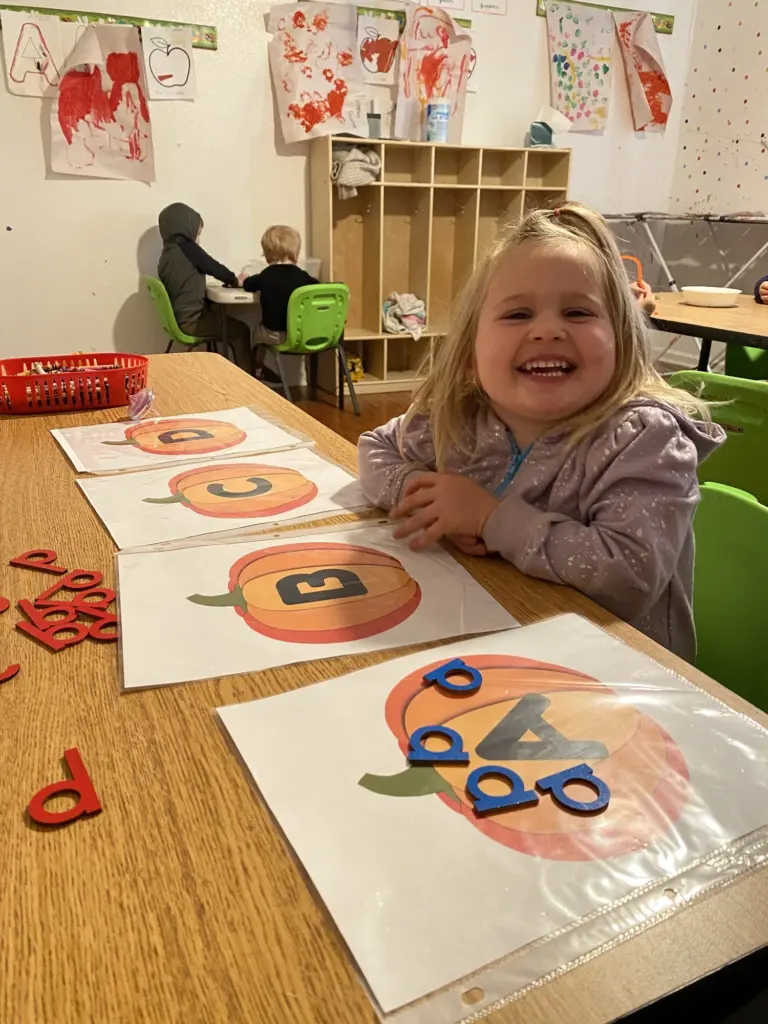 A smiling girl works on a pumpkin letter sheet with red magnetic letters