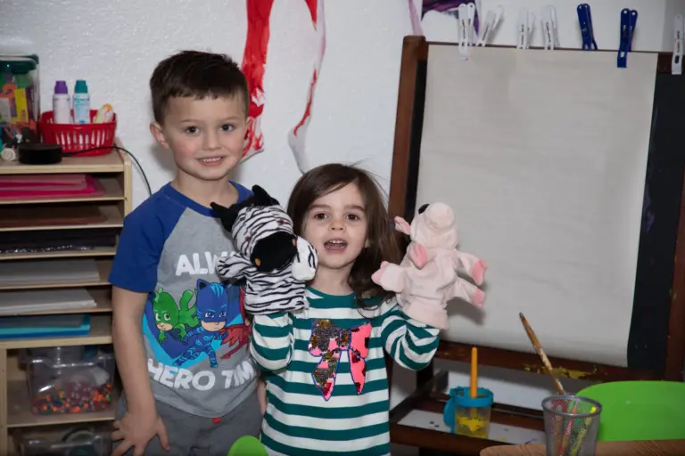 Two children grin at the camera holding a zebra puppet and a pink pig puppet in the classroom