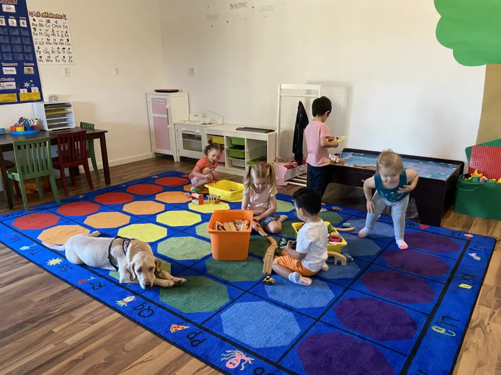 Children play on the big rainbow-dot rug with toys and a sleepy Labrador