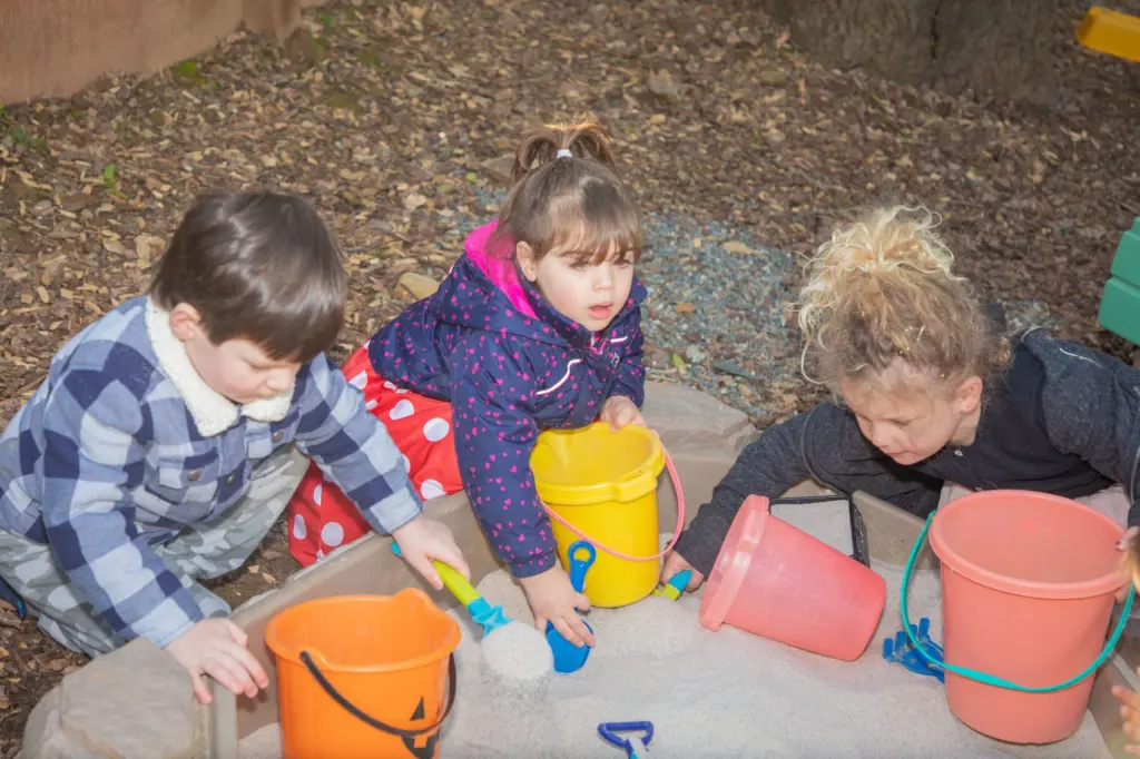 Three children dig in the sandbox with orange and yellow buckets