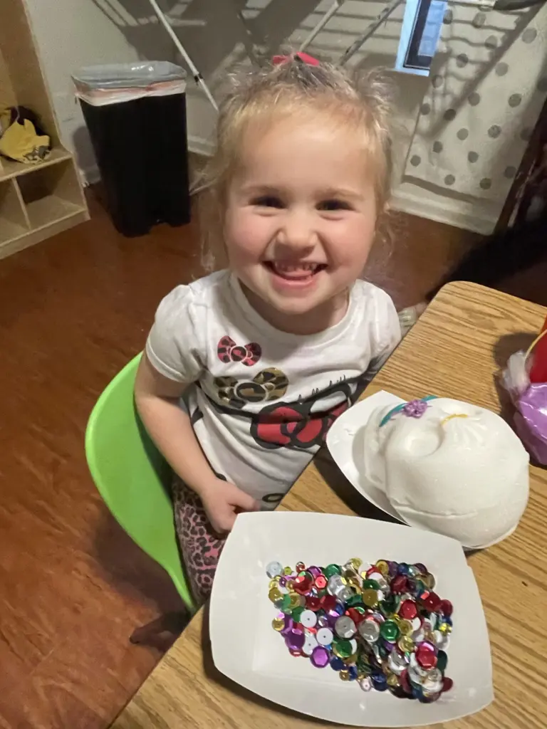 A beaming girl decorates a white mask with sequins at her table