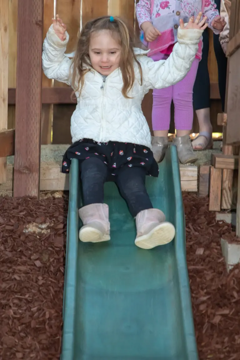 A girl in a white jacket throws her arms up as she slides down the green slide