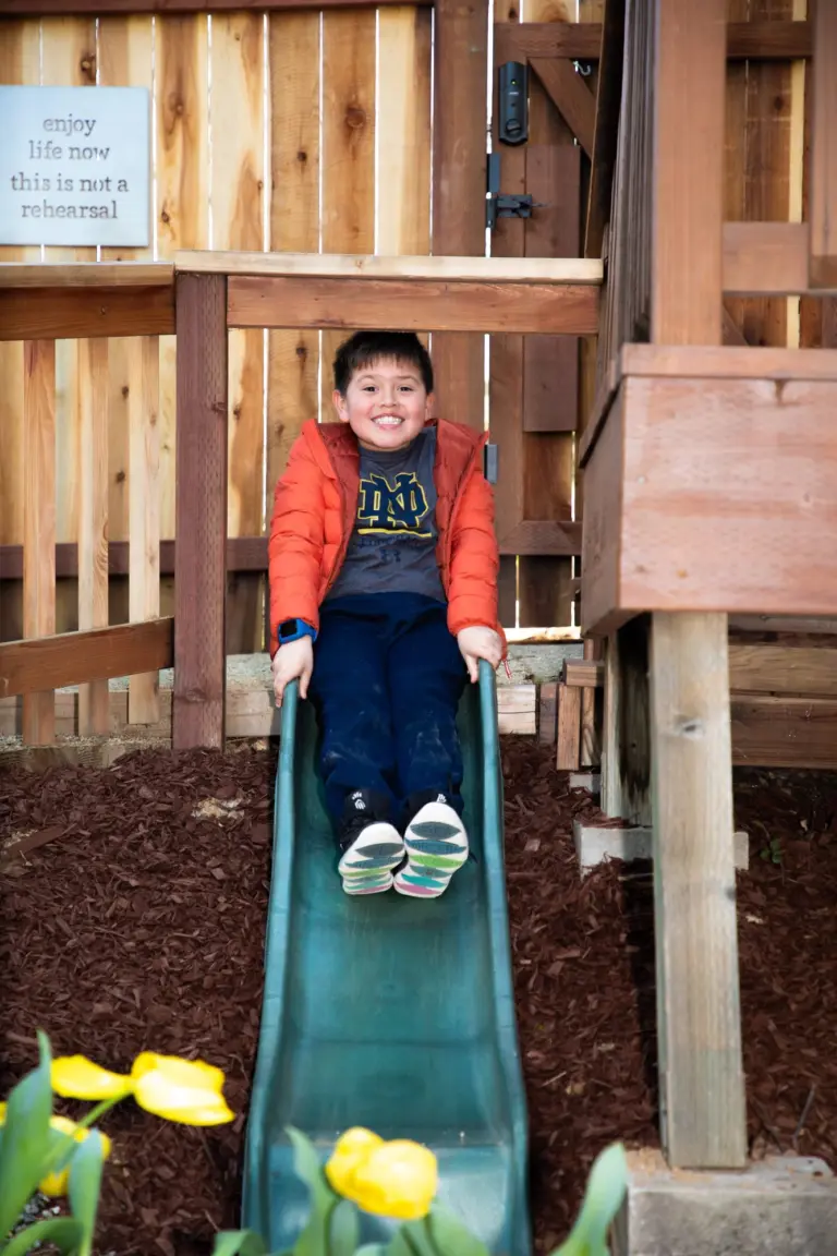 A smiling boy zooms down a green slide in the garden