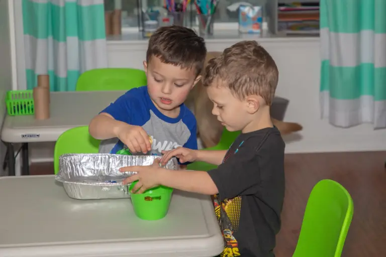 Two boys scoop and sort small green objects from a foil tray at a classroom table