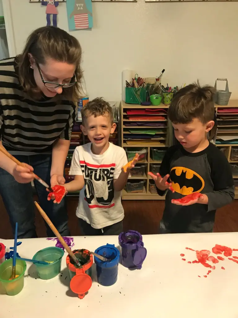 A teacher helps two boys dip their brushes into pots of paint