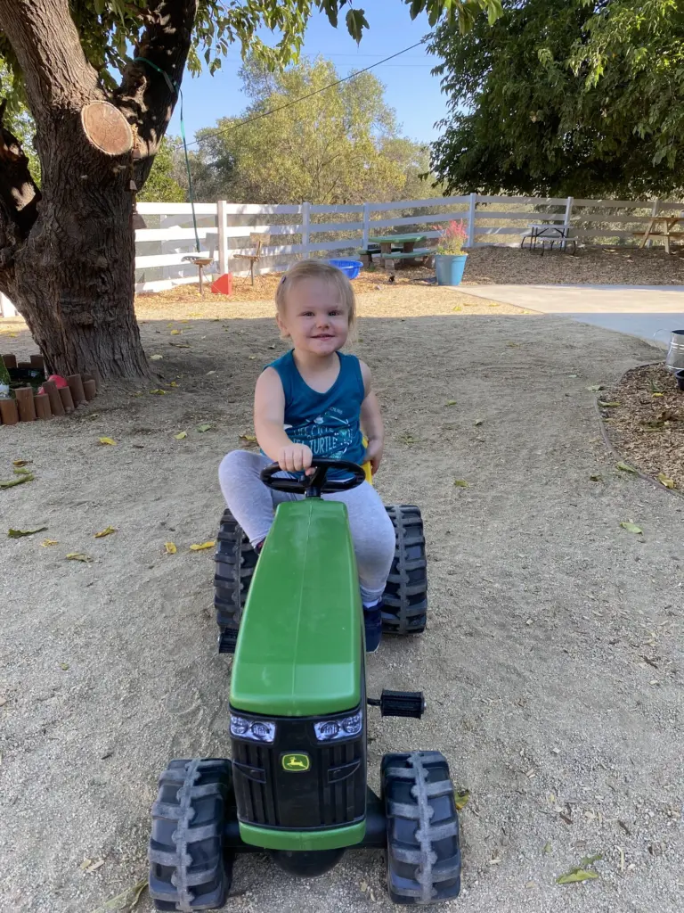 A toddler drives a little green ride-on tractor across the dirt yard under the oaks