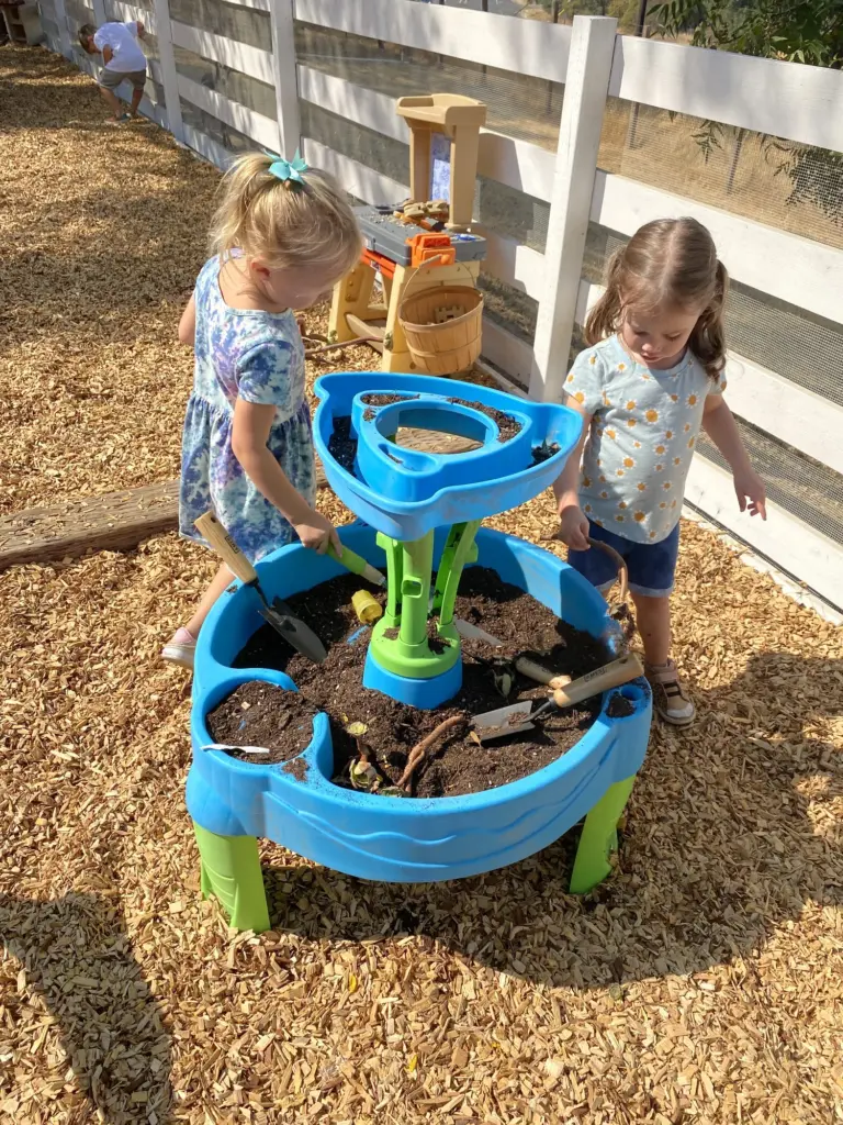 Two girls play at a bright blue water table next to the white farm fence
