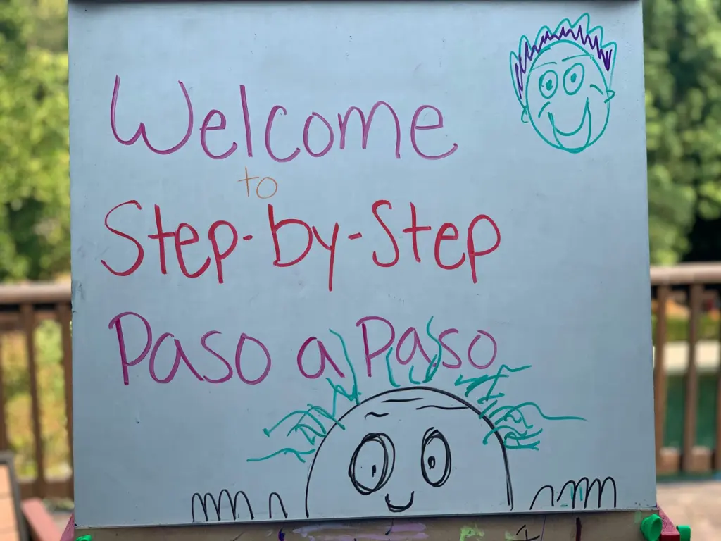 A hand-drawn sign in pink and blue marker that says Welcome to Step-by-Step Paso a Paso, with a smiling sun